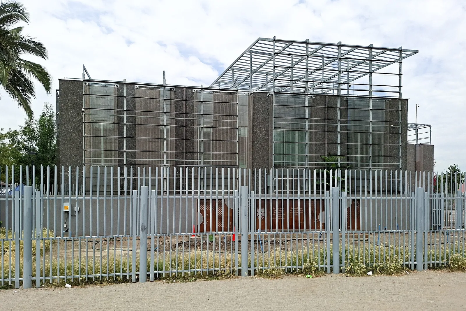 Edificio Consistorial de Huechuraba, Santiago, tras una reja metálica gris, con estructura de pérgola metálica en el techo.