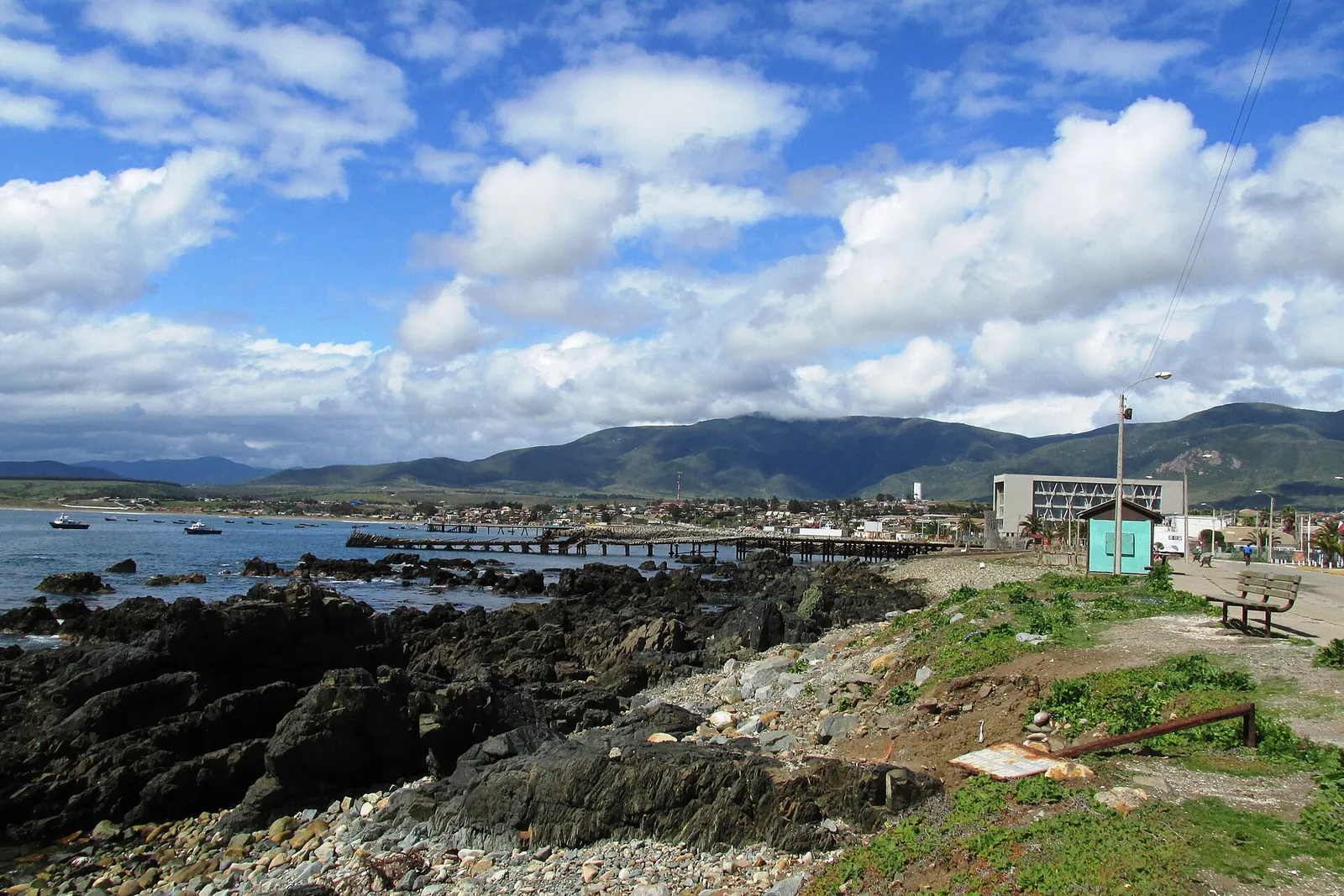 Costa rocosa de Los Vilos, Región de Coquimbo, con un muelle de madera, botes en la bahía y cerros verdes al fondo.