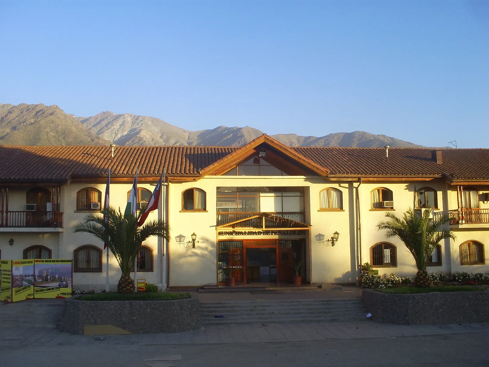Fachada del edificio de la Municipalidad de Peñalolén, Santiago, con techo de tejas, banderas en la entrada y la cordillera al fondo.