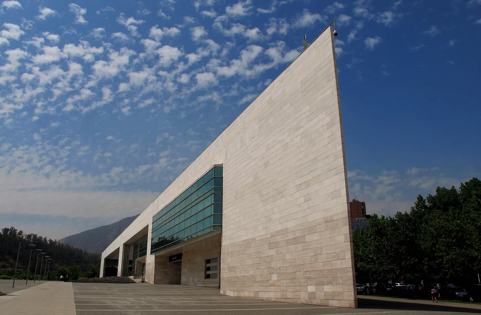 Edificio de la Municipalidad de Vitacura, Santiago: muro de piedra en forma de cuña y fachada vidriada, bajo un cielo con nubes.