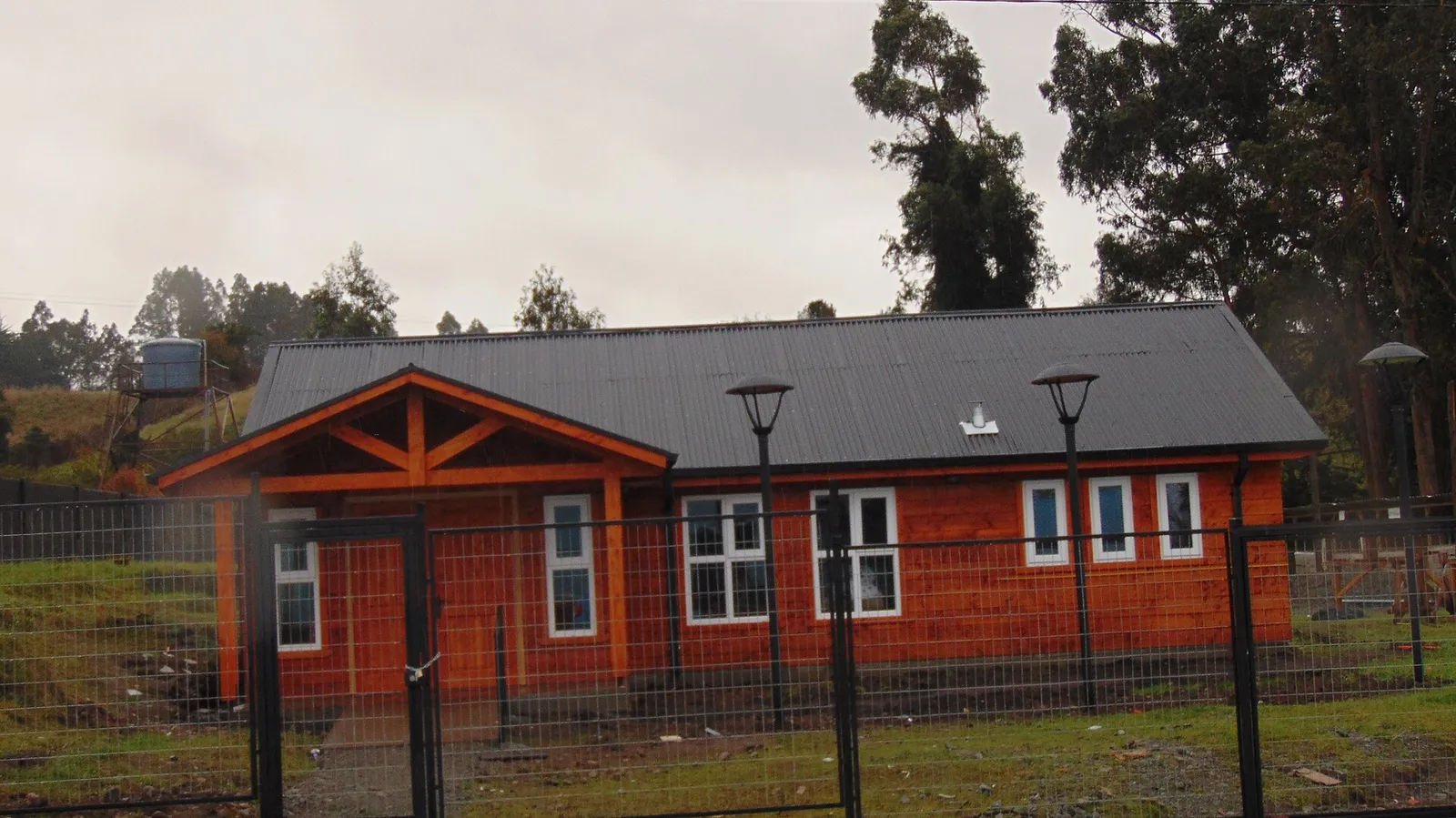 Sede vecinal de madera pintada de rojo, con techo gris y reja perimetral, en el sector Los Bajos de Frutillar, Región de Los Lagos.