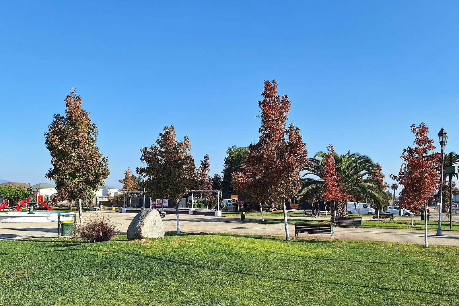 Plaza de Armas de Padre Hurtado, Santiago: prado verde, árboles de follaje rojizo, una palmera y bancas bajo un cielo despejado.