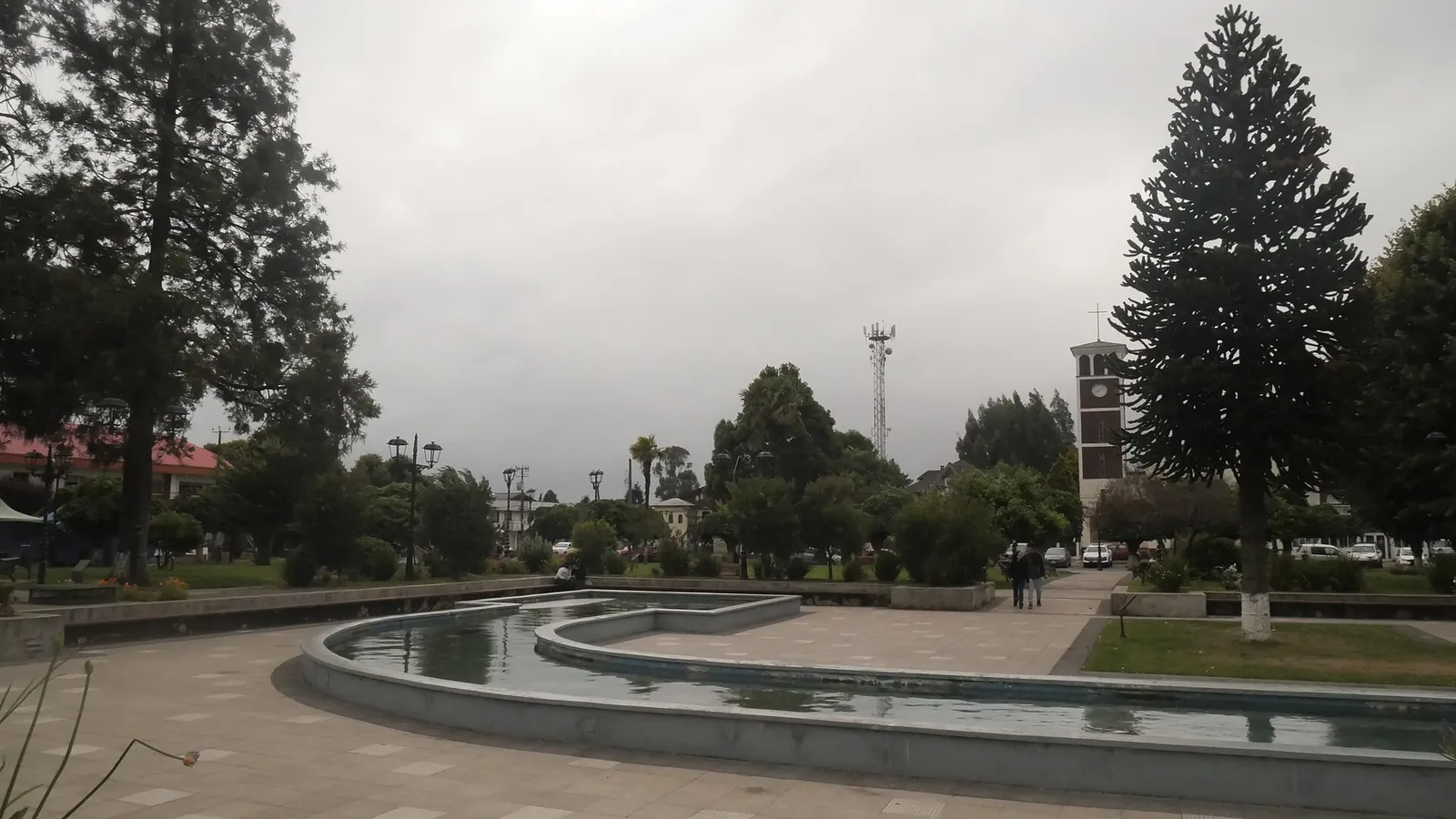 Plaza de Río Bueno, Región de Los Ríos: una pileta con forma curva, araucarias y una torre de iglesia al fondo, bajo un cielo nublado.