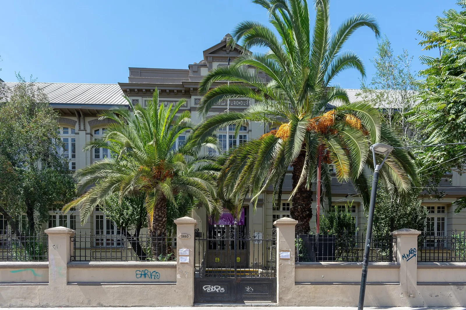 Fachada de la Escuela República El Líbano, en Santiago, con dos palmeras frente al edificio y una reja sobre un muro bajo.