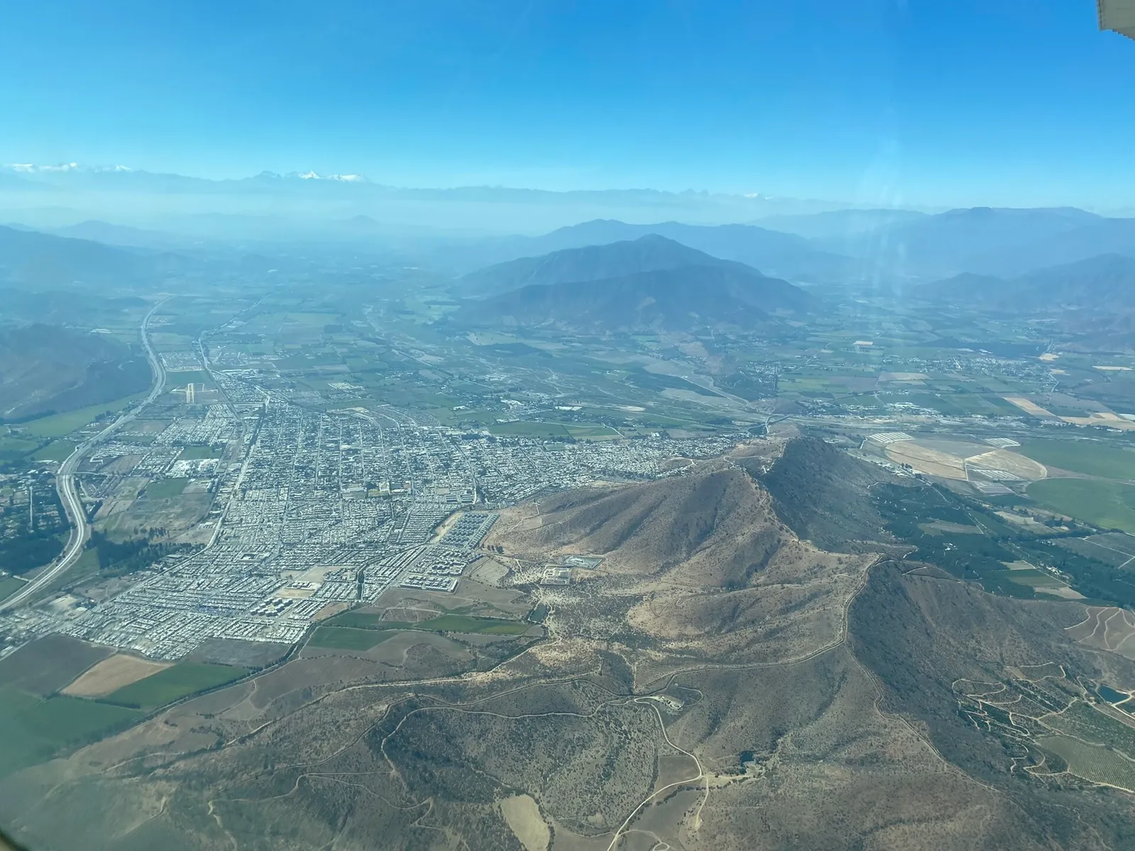Vista aérea de la ciudad de Melipilla y sus alrededores agrícolas, con cerros en primer plano y la cordillera al fondo.