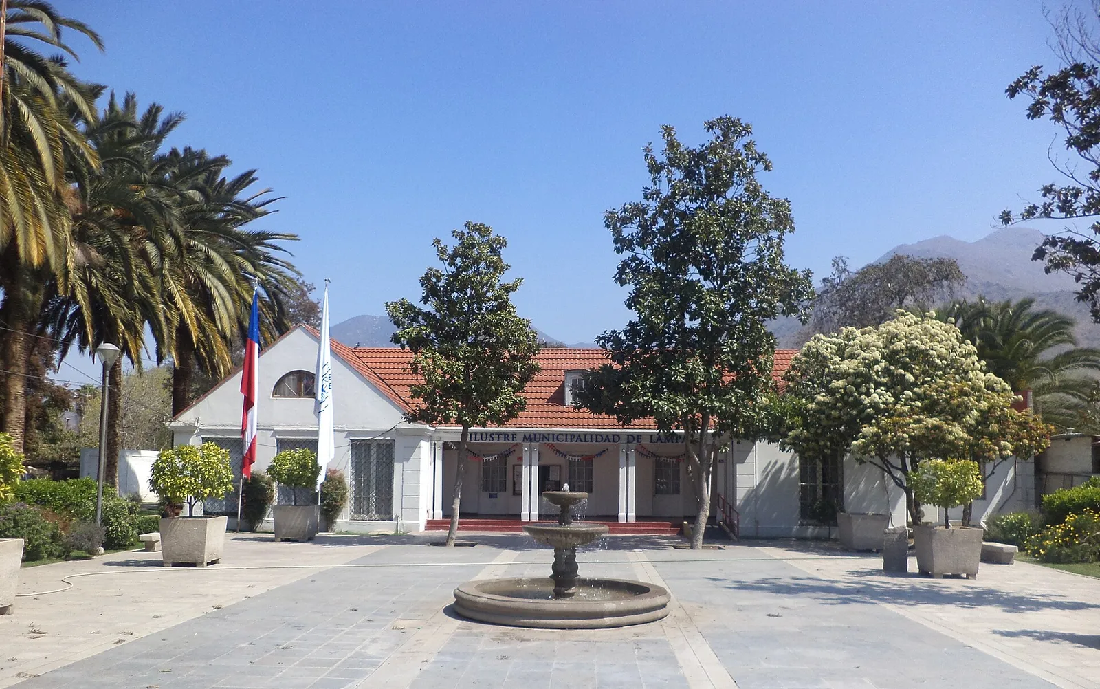 Edificio de la Ilustre Municipalidad de Lampa, de un piso, con una pileta en la explanada, palmeras y cerros al fondo.
