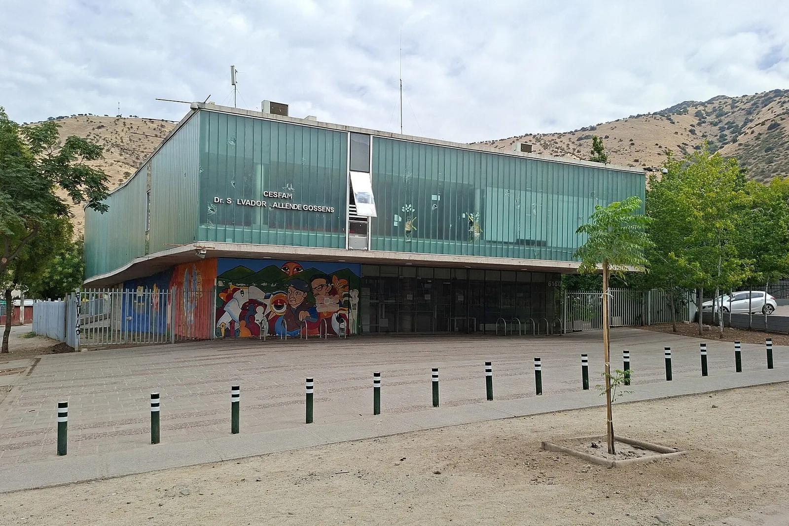Edificio de vidrio verde del Cesfam Dr. Salvador Allende Gossens, en Huechuraba, Santiago, con un mural en la base y cerros al fondo.
