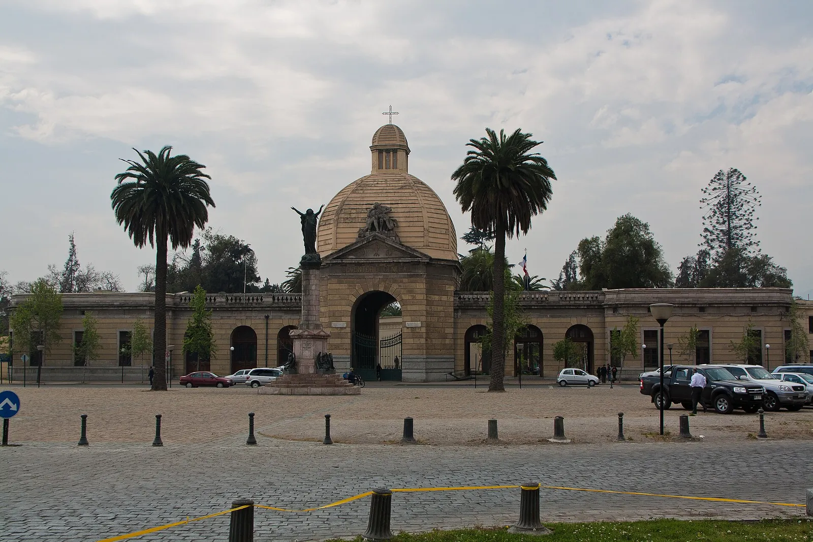Pórtico de acceso del Cementerio General, en Recoleta, Santiago, con su cúpula, dos palmeras y la explanada de estacionamiento delante.