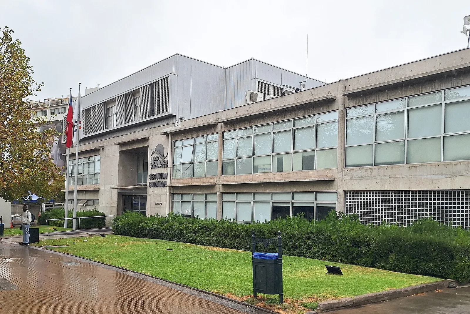 Edificio de la Dirección de Seguridad Ciudadana de Las Condes, Santiago, con prado y banderas frente a la entrada, en un día nublado.