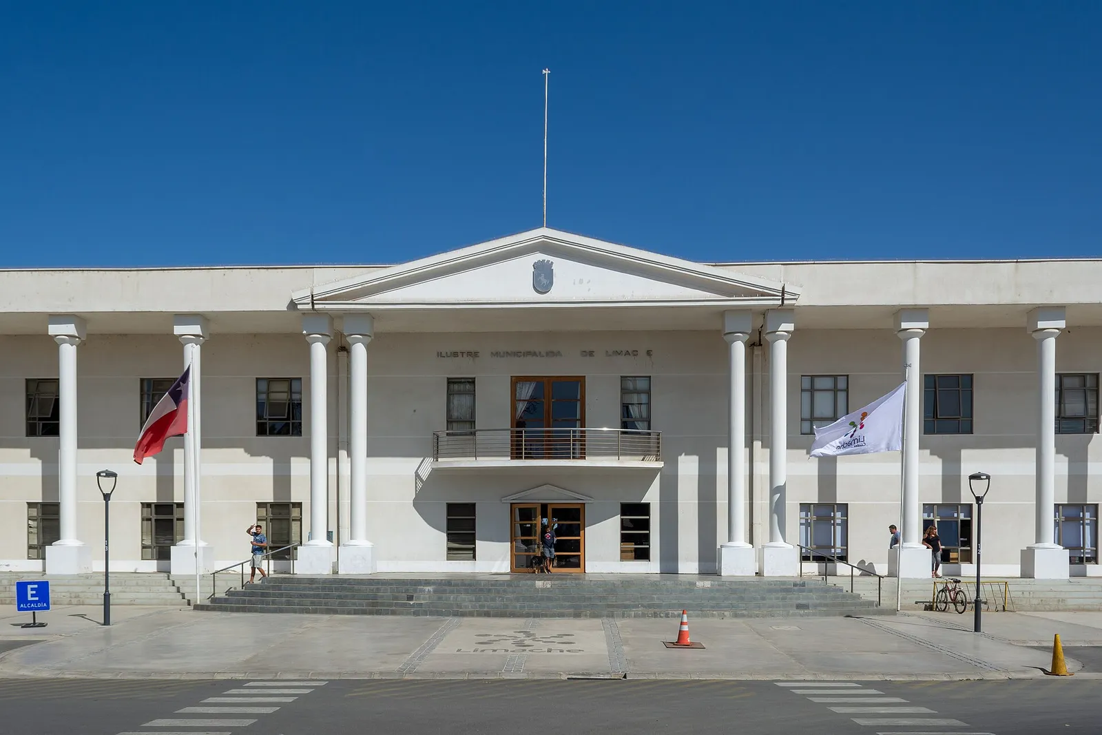 Edificio Consistorial de Limache, Región de Valparaíso: fachada blanca de dos pisos con columnas, frontón, balcón central, banderas y escalinata, bajo cielo despejado.